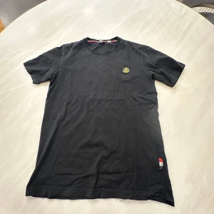 Svart Moncler t-shirt  - Svart t-shirt från Moncler med klassisk passform och rund hals. Liten Moncler-logga på bröstet och en detalj i rött, vitt och blått nedtill. Tillverkad i mjuk bomull, perfekt för en clean och stilren look.