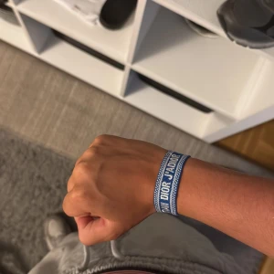 Blått Christian Dior armband - Snyggt blått armband från Christian Dior med vit brodyr och text. Armbandet har justerbara tofsar i ändarna och är gjort i ett mjukt tygmaterial. Perfekt accessoar för att lyfta din outfit med en exklusiv touch.