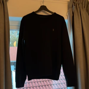 Svart sweatshirt från Polo Ralph Lauren - Svart sweatshirt från Polo Ralph Lauren med klassisk rund hals och diskret broderad logga på bröstet. Tröjan har lång ärm och är tillverkad i mjuk bomullsmix som känns skön mot huden. Perfekt för en clean och stilren look. Tröjan är nästan aldrig använd. Nypris: 1800kr