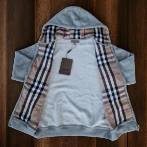 Grå hoodie från Burberry med dragkedja - Snygg grå hoodie från Burberry i 100% bomull med klassiskt rutigt foder i huvan och insidan. Tröjan har dragkedja, snörning i huvan och broderad Burberry-logga på bröstet. Perfekt för dig som vill ha en stilren och lyxig look.