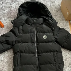 Svart pufferjacka från Trapstar M - Säljer en svart pufferjacka från Trapstar med stor huva och logga på bröstet. Jackan har broderad Trapstar-text på ryggen och dragkedja framtill. Fylld för att hålla dig varm och har en clean streetwear vibe. Perfekt för kalla dagar.