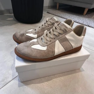 Maison Margiela Replica sneakers beige - Maison Margiela Replica sneakers i beige och vitt med klassisk retrodesign. Ovandel i mocka och skinn, bruna gummisulor och snörning. Skick 8/10, någon missfärgning som säkert gå bort med en tvätt. Storlek 44, allt og följer med men kvitto saknas. Äktahetsbevis saknas därav priset. Pris går att diskutera✅