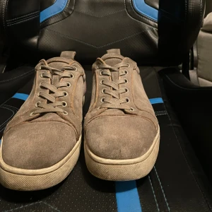 Bruna mockasneakers från Christian Louboutin - Säljer ett par gråa sneakers från Christian Louboutin i mocka med klassisk röd sula och vita kanter. Skorna har snörning och rund tå, samt en stilren och enkel design som passar till många outfits. Kommer med originalkartong. Har fått ett hål i hälen men de är fixat och syns inte vid användning