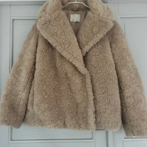 Beige teddyjacka med bred krage - Supermysig teddyjacka i beige med bred krage och långärmad design. Jackan har en fluffig och mjuk yta som ger en chill vibe och är perfekt för kalla dagar. Modellen är loose fit och har en klassisk look utan synliga detaljer som knappar eller dragkedja.