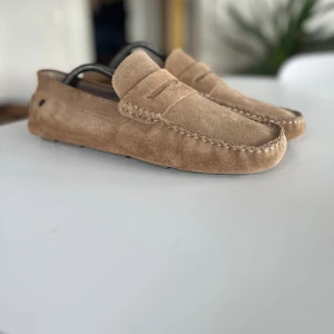 Beige mockaloafers i storlek 43 - Helt nya loafers från Jack and Jones i beige mocka med klassisk pennyloafers-design. Skorna har en mjuk ovandel, dekorativa sömmar och en flexibel gummisula med mönster för extra grepp. Perfekta för en stilren och avslappnad look.