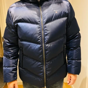 Mörkblå pufferjacka från Gant, L - Snygg mörkblå pufferjacka från Gant i storlek L. Jackan har quiltad design, dragkedja framtill och hög krage med luva. Perfekt för kalla dagar med sin vadderade känsla och klassiska look. Gant-logga på bröstet och praktiska fickor med dragkedja. Jackan är som ny! 
