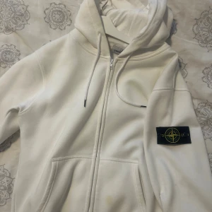 Vit hoodie från Stone Island - Vit hoodie från Stone Island med dragkedja framtill och klassisk logga på vänster ärm. Tröjan har huva med snörning och två fickor framtill. Mjuk och skön, perfekt för chill dagar. Enkel och stilren design som passar till det mesta.