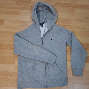 Grå hoodie från Polo Ralph Lauren XL - Klassisk grå hoodie från Polo Ralph Lauren i storlek XL=160cm . Tröjan har dragkedja, huva och fickor framtill. Broderad marinblå logga på bröstet. Mjuk och skön bomullsblandning, perfekt för chill dagar.