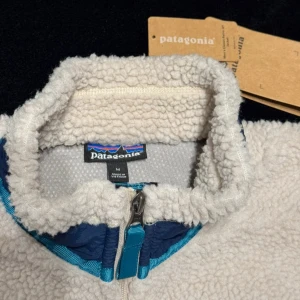  Patagonia jacka - Säljer en jacka från Patagonia i storlek M. Jackan har mjuk och fluffig fleece, blå detaljer vid dragkedjor och krage samt en praktisk bröstficka med zip. Perfekt för dig som gillar chill och varm stil under kyliga dagar.