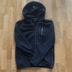 Svart hoodie från Sail Racing med dragkedja - Svart hoodie från Sail Racing med hel dragkedja, broderad logga på bröstet och en extra bröstficka med dragkedja. Mjuk insida och huva med broderad detalj. Perfekt för kyliga dagar och snygg till jeans eller joggers.