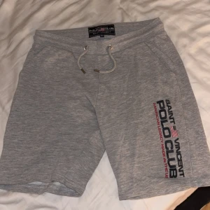 Grå shorts från Polo Club Saint Vincent - Snygga grå shorts från Polo Club Saint Vincent med tryck på benet och bakfickan. De har elastisk midja med snörning och är gjorda i mjuk bomull, perfekta för chill dagar. Klassisk loose passform och neutralt utseende som funkar till det mesta.