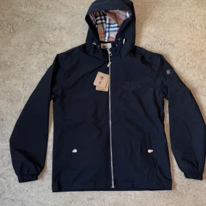 Svart vindjacka från Burberry M - Svart vindjacka från Burberry med klassiskt rutigt foder i huvan, dragkedja framtill och två fickor med tryckknappar. Jackan har broderad Burberry-text på bröstet och TB-logga på ärmen. Snygg och stilren modell med justerbar huva.