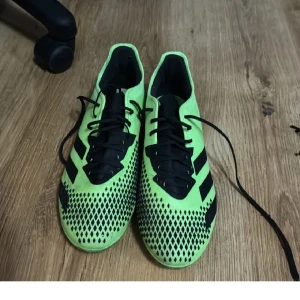 Gröna Adidas fotbollsskor med snörning - Säljer ett par grön-svarta Adidas fotbollsskor med klassiska svarta ränder på sidorna och svart snörning. Ovandelen har ett mönster med svarta rutor och skon är designad för att ge bra grepp på planen. Perfekta för dig som vill sticka ut på matchen.