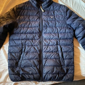 Mörkblå pufferjacka från GANT - Säljer en mörkblå pufferjacka från GANT med horisontella sömmar och dragkedja framtill. Jackan har två snedställda fickor och står för en clean och sportig look. Perfekt för kyliga dagar och enkel att matcha med det mesta.