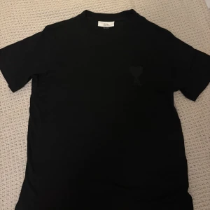 Svart t-shirt från Ami Paris - Svart t-shirt från Ami Paris med diskret broderad logga med hjärta och A på bröstet. Klassisk passform och rund halsringning. Tillverkad i mjuk bomull för en skön känsla. Perfekt för en clean och stilren look. Använda fåtal gånger men tvättad en gång därför är priset justerat efter det!