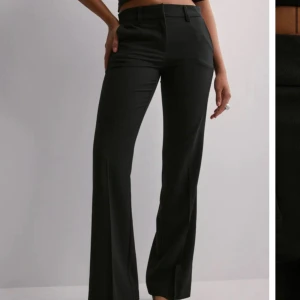 Svarta kostymbyxor flare Nelly - Snygga svarta kostymbyxor med hög midja och wide fit. Byxorna har klassisk rak design, pressveck och stängs med dragkedja och knapp framtill. Perfekta för en stilren och trendig look.