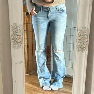 Ljusblå bootcut jeans med slitningar - Säljer ett par ljusblå jeans med bootcut passform och slitna detaljer vid knäna. Jeansen har klassisk femficksdesign, knapp och dragkedja fram samt fickor med lock bak. Materialet är mjukt denim och midjan är normal.