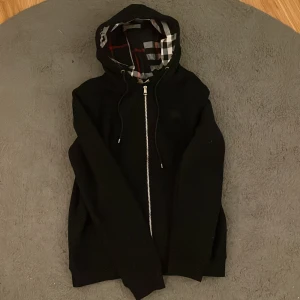 Svart Burberry hoodie med dragkedja XL - Svart hoodie från Burberry i 100% bomull med klassiskt rutigt foder i huvan. Tröjan har dragkedja framtill, snörning vid huvan och en clean look. Perfekt för dig som gillar stilrena märkesplagg med en twist.