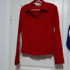 Röd skjorta från Åhléns, storlek 36 - Klassisk röd skjorta från Åhléns i storlek 36. Skjortan har lång ärm, knappar framtill och en snygg krage. Perfekt för dig som vill sticka ut med en färgstark look. Materialet känns mjukt och lätt, troligen bomull eller polyester.