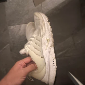Nike Presto sneakers i vitt mesh - Nike Presto sneakers i helvitt med mesh-överdel och syntetdetaljer. Skorna har rund tå, snörning och en platt sula med ventilerande hål. Swoosh-logga på sidan och mjuk innersula för extra komfort. Perfekt för dig som gillar stilrena och sportiga sneakers.