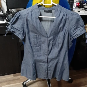 Blå kortärmad skjorta från Lindex - Snygg blå skjorta från Lindex i luftig bomull med korta ärmar och knytband. Skjortan har två bröstfickor med knappar och en avslappnad passform. Perfekt till jeans eller kjol för en chill look.