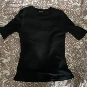 Svart ribbad topp från Monki - Säljer en svart ribbad topp från Monki i storlek S. Toppen har rund halsringning och korta ärmar, med en slim passform som sitter snyggt på kroppen. Materialet är mjukt och lätt, perfekt för dig som gillar stilrena och enkla plagg.