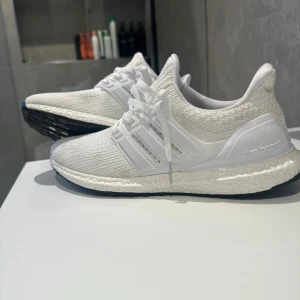 Vita Adidas Ultraboost sneakers - Säljer ett par fräscha vita Adidas Ultraboost sneakers med stickad ovandel och klassiska tre ränder på sidan. Skorna har snörning och en bekväm, responsiv sula som ger grym komfort. Perfekta för dig som gillar stilrena och sportiga skor. 