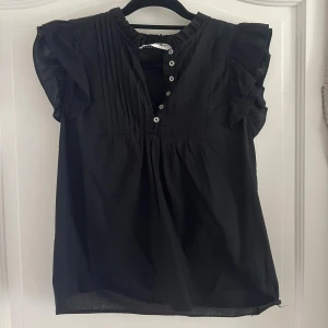 Svart blus med volang från Co'couture - Svart blus från Co'couture i storlek S. Blusen har korta volangärmar, knappar framtill och veckade detaljer vid bröstet.