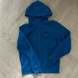 Blå hoodie från Peak Performance - Säljer en blå hoodie från Peak Performance med dragkedja och huva. Tröjan har tryckt logga på bröstet och praktiska fickor framtill. Perfekt för en avslappnad stil och enkel att matcha med jeans eller träningsbyxor.