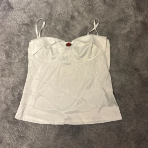 Vit satin topp med rosdetalj - Snygg vit topp i satin med smala axelband och en liten röd rosdetalj framtill. Toppen har formade kupor och en lätt glansig finish som ger en lyxig känsla. 