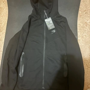 Nike tech fleece kofta - Svart hoodie från Nike med hel dragkedja och huva. Tröjan har diskret Nike-logga på bröstet, två fickor med dragkedja och är tillverkad i ett mjukt funktionsmaterial. Perfekt för träning eller chill. Snygg och enkel design som passar till allt.