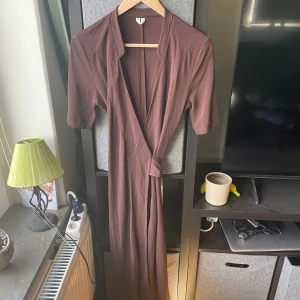 Brun omlottklänning med knyt XS - Snygg lång brun omlottklänning i mjukt material med korta ärmar och knyt i midjan. Stilren och enkel design som ger en chill vibe, perfekt för dig som gillar minimalistiskt mode. Passar dig som vill ha en klänning med lite extra längd och skön passform.