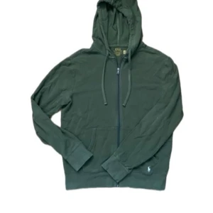 Grön hoodie från Polo Ralph Lauren - Mörkgrön hoodie från Polo Ralph Lauren med dragkedja och huva. Klassisk broderad logga i vitt på fickan. Tröjan har långa ärmar, ribbade muddar och snörning i huvan. Perfekt för chill dagar eller när du vill vara extra bekväm.