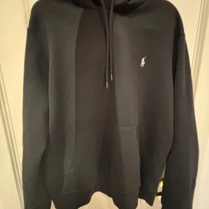 Svart hoodie från Polo Ralph Lauren - Svart hoodie från Polo Ralph Lauren med klassisk logga broderad på bröstet. Tröjan har huva med snörning, stor magficka och är tillverkad i en mjuk blandning av bomull och polyester. Perfekt för chill dagar eller när du vill ha en clean look.