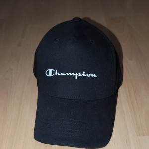 Svart keps från Champion - Säljer en svart keps från Champion med klassisk böjd skärm och justerbar rem baktill. Kepsen har vit Champion-logga broderad framtill och är tillverkad i bomull. Insidan har detaljer med Champion-tryck längs sömmarna. Perfekt accessoar för en sportig look.