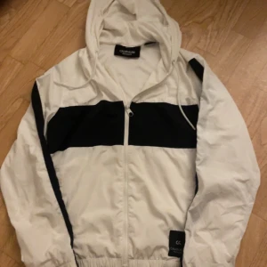 Vit och svart vindjacka Calvin Klein - Snygg vit vindjacka från Calvin Klein med svarta detaljer över bröstet och längs ärmarna. Jackan har huva med dragsko, hel dragkedja framtill och elastiska muddar. Perfekt för en sportig och clean look. Köpt i zalando för 600kr och inte använt mycket! Pris kan diskuteras!