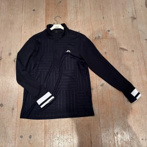 Blå half zip tröja från J.Lindeberg - Svart långärmad tröja från J.Lindeberg med diskret geometriskt mönster och vitt JL-logga på bröstet. Tröjan har half zip, ståkrage och vita detaljer vid ärmsluten. Perfekt för dig som gillar stilren sportig look.