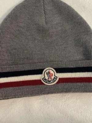 Moncler mössa - Säljer en snygg grå Moncler mössa med stickad struktur och klassiska Moncler-loggan framtill. Mössan har en randig detalj i svart, vitt och rött längs kanten som ger en sportig vibe. Perfekt för kalla dagar och enkel att matcha med jacka eller hoodie.