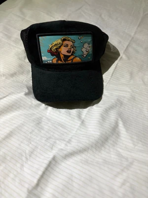 Svart keps från Revenite - Säljer en svart keps från Revenite med coolt pop art-tryck framtill som föreställer en tecknad person i färgglada toner. Kepsen har mesh på baksidan och justerbar snapback. Perfekt accessoar för att lyfta din outfit.