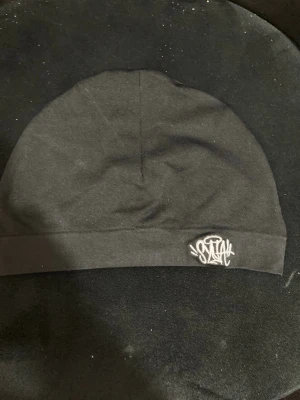 Svart mössa från syna world - Svart mössa med stilren design och broderad sunna wrold-logga i vitt framtill. Mössan är mjuk och stretchig, perfekt för att hålla värmen. Passar dig som gillar enkel och urban stil.