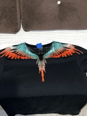 Svart t-shirt med färgglada fjädrar - Säljer en svart t-shirt från Marcelo Burlon med ett fett tryck av färgglada fjädrar i turkos, orange och grått runt halsen. T-shirten har rund hals och korta ärmar, tillverkad i mjuk bomull. Perfekt för dig som vill sticka ut med en unik design.