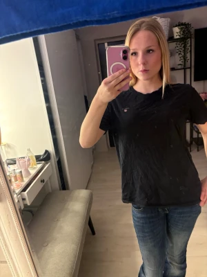 Svart t-shirt från GANT - Svart t-shirt från GANT med liten broderad logga på bröstet. Klassisk rund halsringning och korta ärmar. Tillverkad i mjuk bomull som känns skön mot huden. Perfekt till jeans eller shorts för en enkel och clean look.