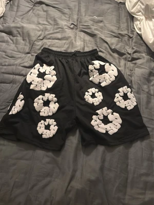 Svarta shorts från Denim Tears med tryck - Svarta shorts från Denim Tears med elastisk midja och snörning. De har stora vita blommönster tryckta över hela plagget, vilket ger en unik och cool vibe. Materialet känns som bomull och passformen är loose för extra komfort.