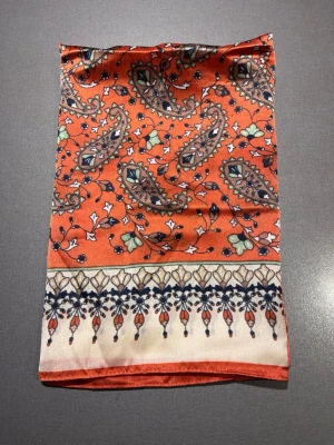 Mönstrad scarf från Åhléns - Scarf från Åhléns i orange med paisley inspirerat mönster och detaljer i beige, svart och grönt. Materialet är mjukt och glansigt, troligen polyester eller siden. Mått: 50x50cm. Mycket bra skick, som ny! Säljs på flera sidor, först till kvarn. 
