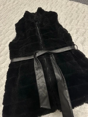 Svart pälsväst med bälte - Säljer en svart pälsväst med mjuk och fluffig yta. Västen har ett snyggt bälte i skinnimitation som knyts i midjan. Storlek S från amisu. 