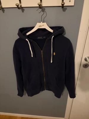 Marinblå hoodie från Polo Ralph Lauren - Snygg marinblå hoodie från Polo Ralph Lauren med gul broderad logga på bröstet. Tröjan har huva med vita snören, dragkedja framtill och två fickor. Perfekt för chill dagar och har en klassisk streetstil.