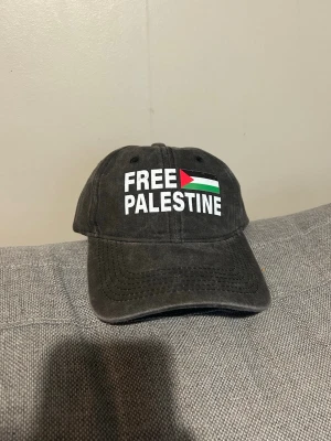 Svart keps med Free Palestine-tryck - Svart keps med böjd skärm och justerbart spänne bak. Framsidan har ett tryck med texten 'FREE PALESTINE' och en flagga i rött, grönt, vitt och svart. Kepsen är tillverkad i bomull och har en klassisk, avslappnad look som passar till streetwear.FREE PALESTINE KEPS - GÅRDA WASHED   Detaljinformation:  Tillverkad av 100 procent bomull One Size Justerbar vid kepsens baksida Gjord av: 100% bomull  