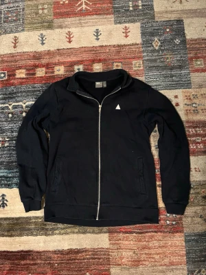 Blå ziptröja från ASOS i bomull - Blå långärmad ziptröja från ASOS i bomull. Tröjan har hel dragkedja framtill, ståkrage och två fickor. En liten vit triangel-logga på bröstet ger en clean look. Perfekt till chill eller när du vill hålla det enkelt.