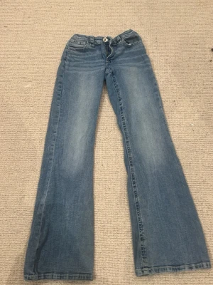 Blå bootcut jeans med detaljer - Säljer ett par blå jeans med bootcut-snitt och snygga detaljer på bakfickorna. Jeansen har klassisk femficksdesign, knapp och dragkedja framtill samt ljusare tvätt på låren. Perfekta för dig som gillar en avslappnad och trendig look.