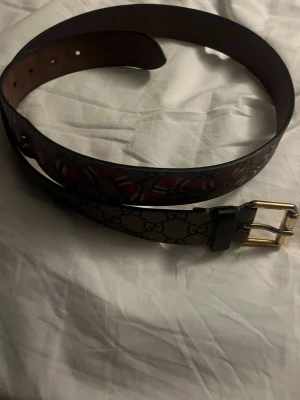 Gucci bälte med GG-mönster och orm - Säljer ett Gucci bälte med klassiskt GG-mönster i beige och brunt, dekorerat med en röd och vit orm längs bandet. Bältet har en guldfärgad metallspänne och är tillverkat i skinn. Perfekt för dig som vill ha en statement-accessoar.